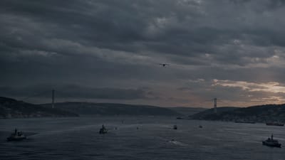 Naval patrol vessels and a small uncrewed aircraft over a calm strait at dusk.