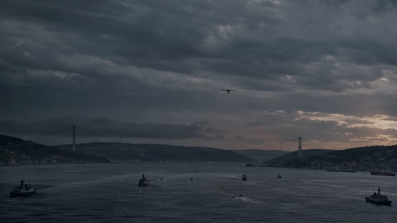 Naval patrol vessels and a small uncrewed aircraft over a calm strait at dusk.